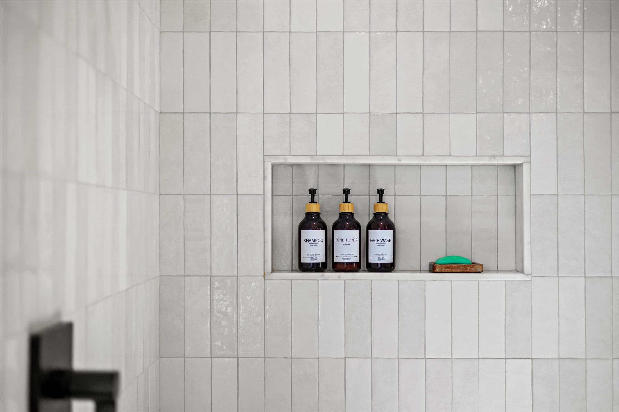 Detail of recessed shower niche with white tile surround, matte black shower valve and handle below, styled with amber product bottles