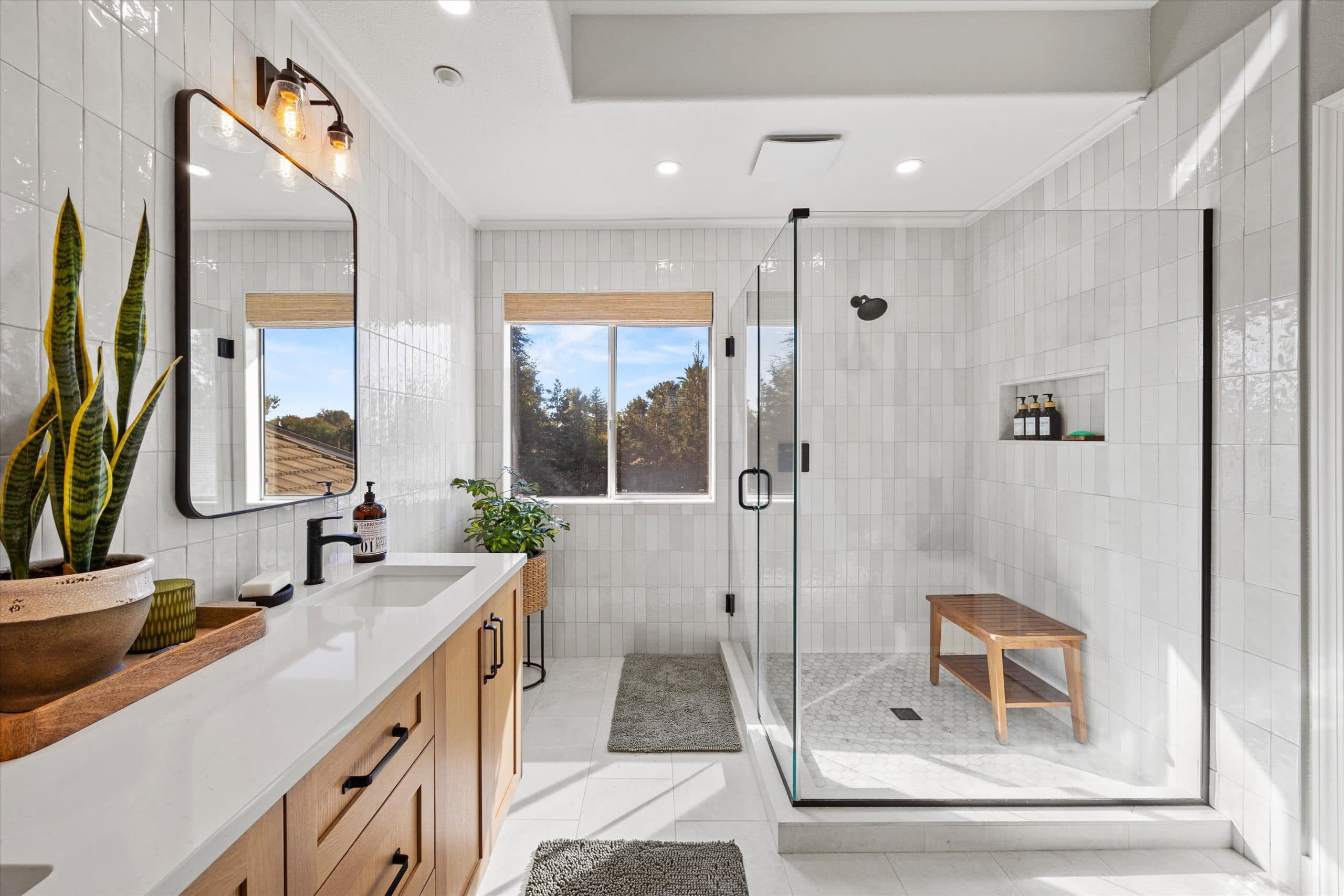 Wide view of remodeled master bathroom showing natural wood double vanity, black-framed glass shower enclosure, teak bench, hexagonal floor tile, window with natural light, and white subway tile walls