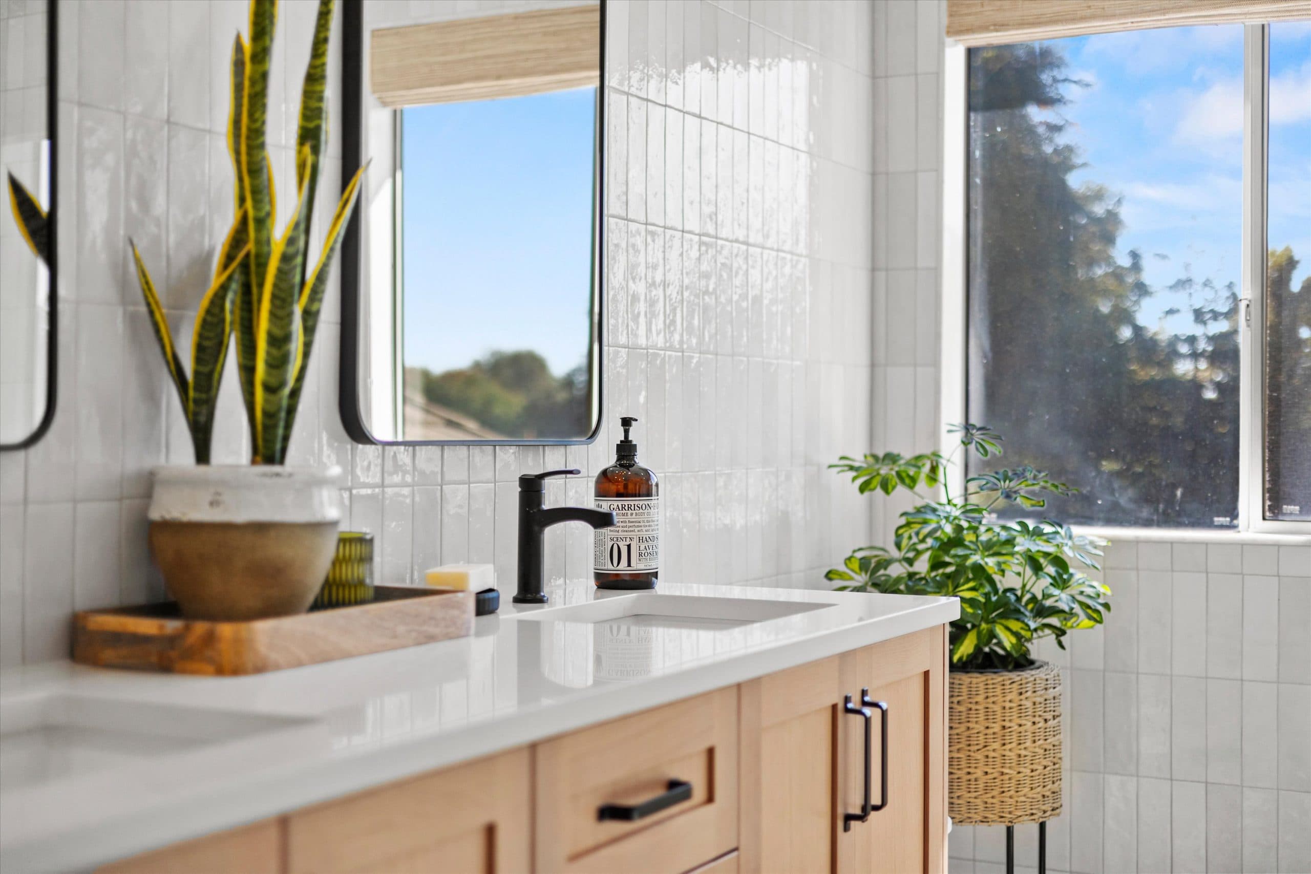 Sink detail showing matte black single-handle faucet on white countertop, natural wood vanity, snake plant in pot, amber soap dispenser, and large window with natural light