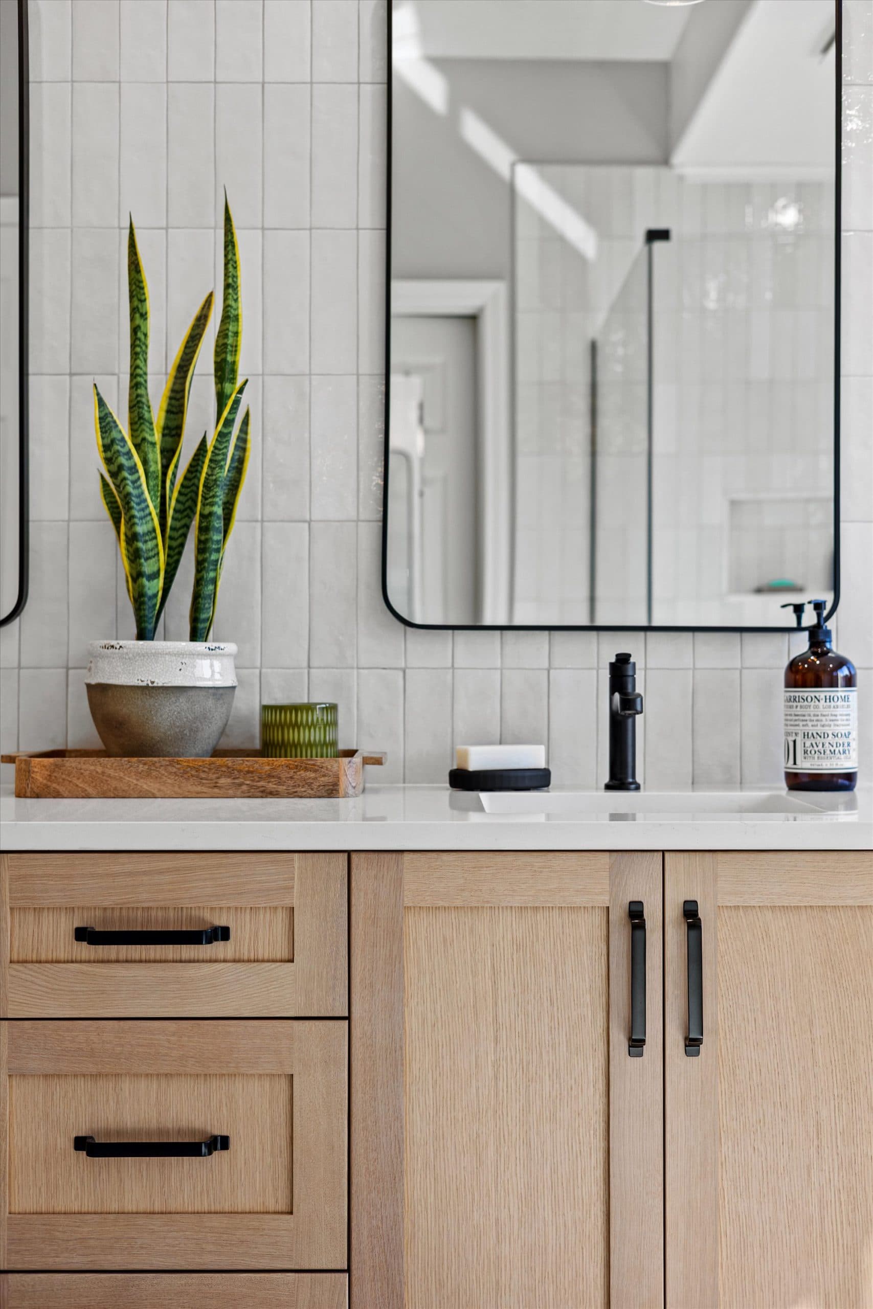 Portrait view of single vanity side featuring black-framed mirror, white subway tile backsplash, snake plant, amber soap bottle, and natural wood cabinet with matte black hardware