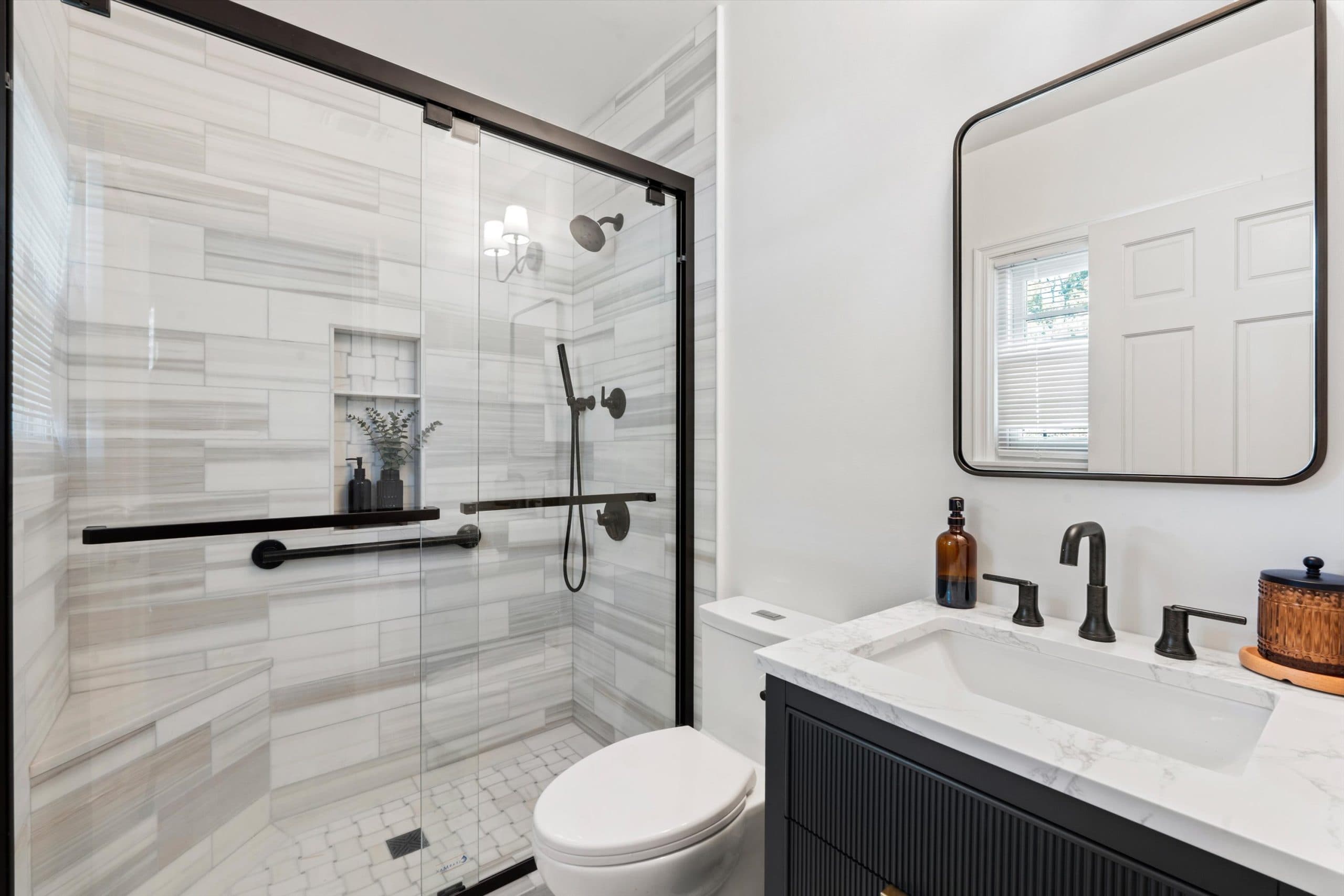 Wide view of remodeled bathroom showing dark navy vanity with white vessel sink, black-framed mirror, and glass-enclosed shower with gray tile and built-in niche
