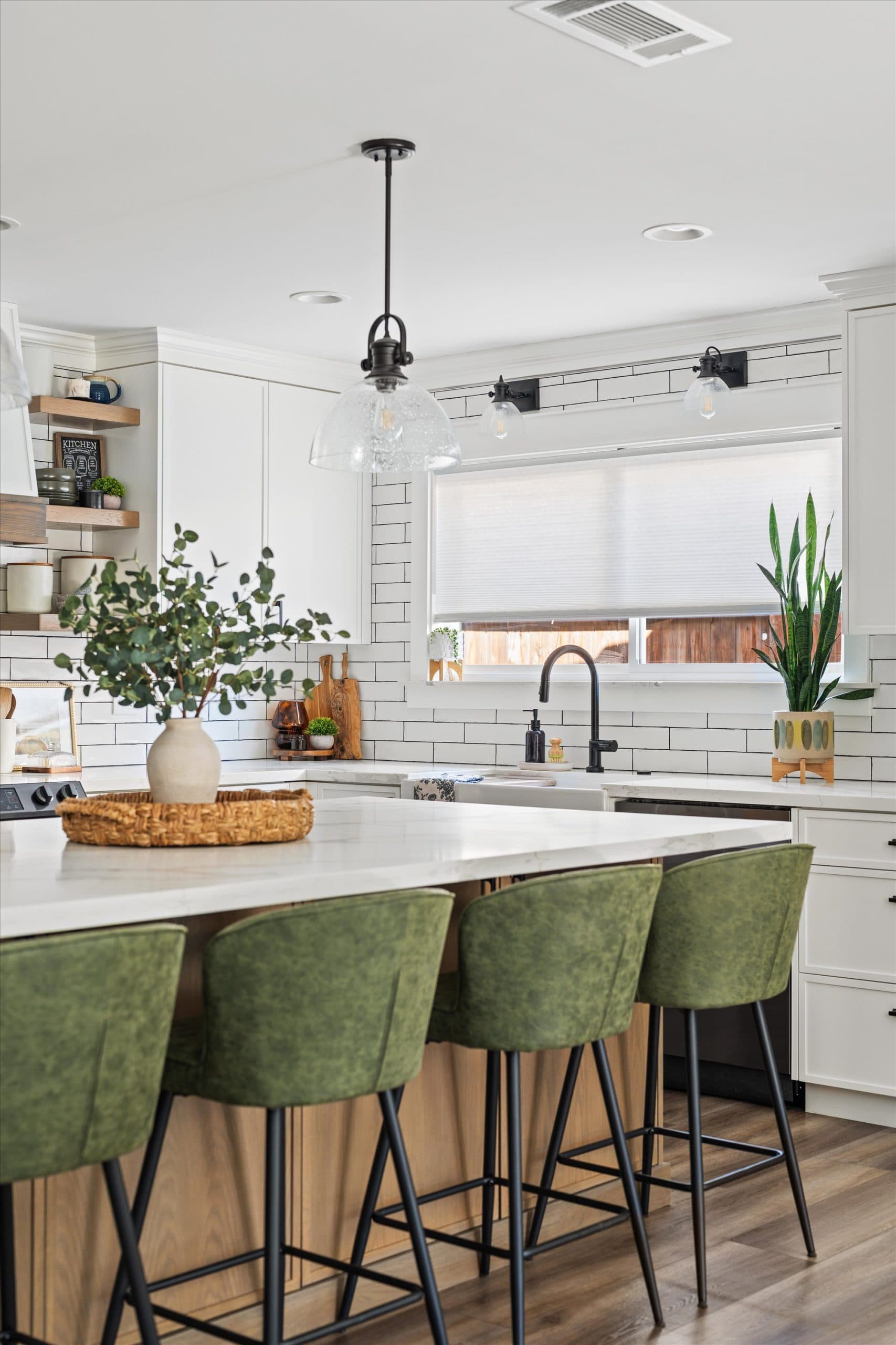 Angled view of kitchen island with green velvet bar stools, black pendant lights, white quartz countertops, and open shelving with plants and kitchen accessories