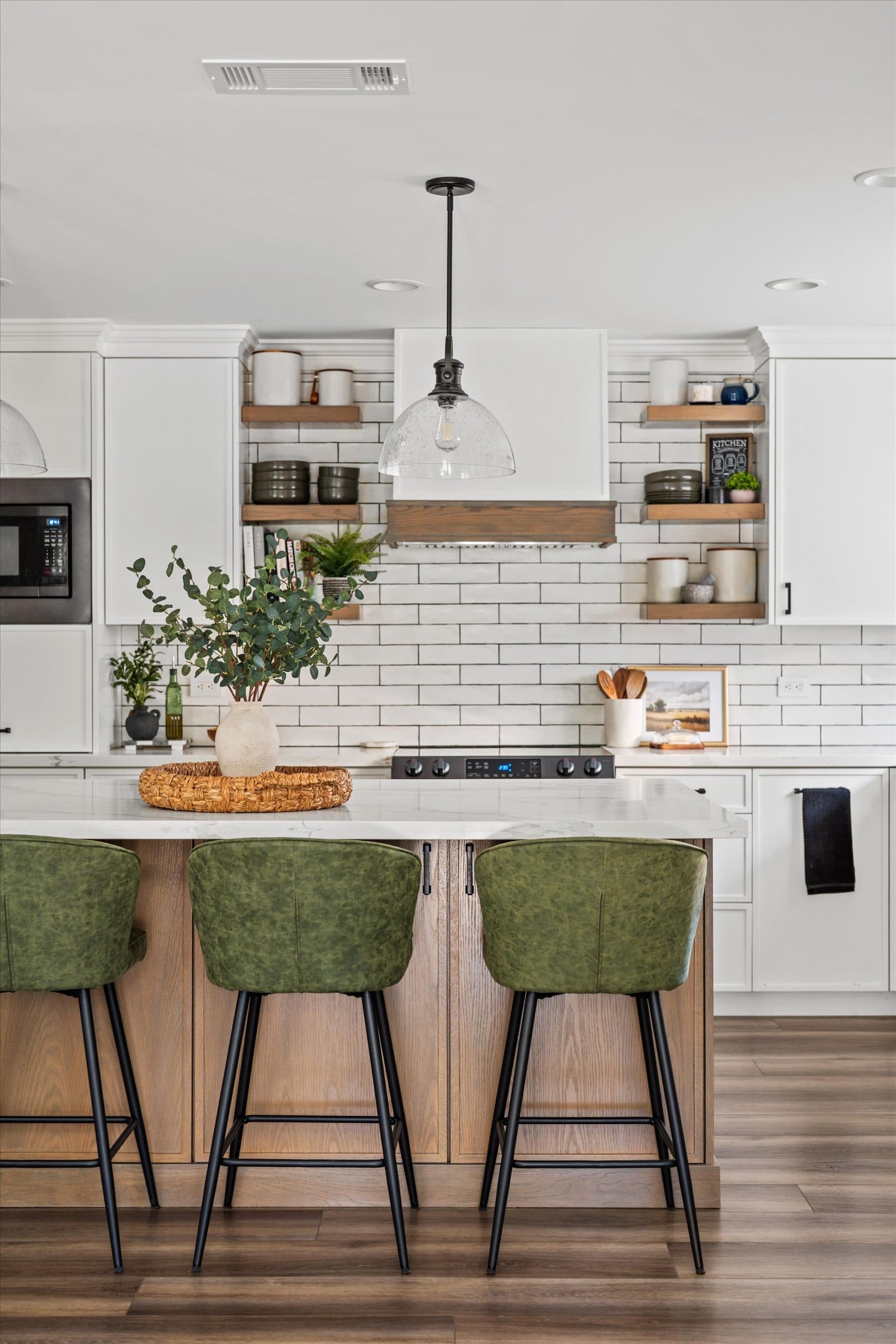 Front view of kitchen island with three green upholstered bar stools, black pendant lighting, white cabinetry, and floating wood shelves in the background