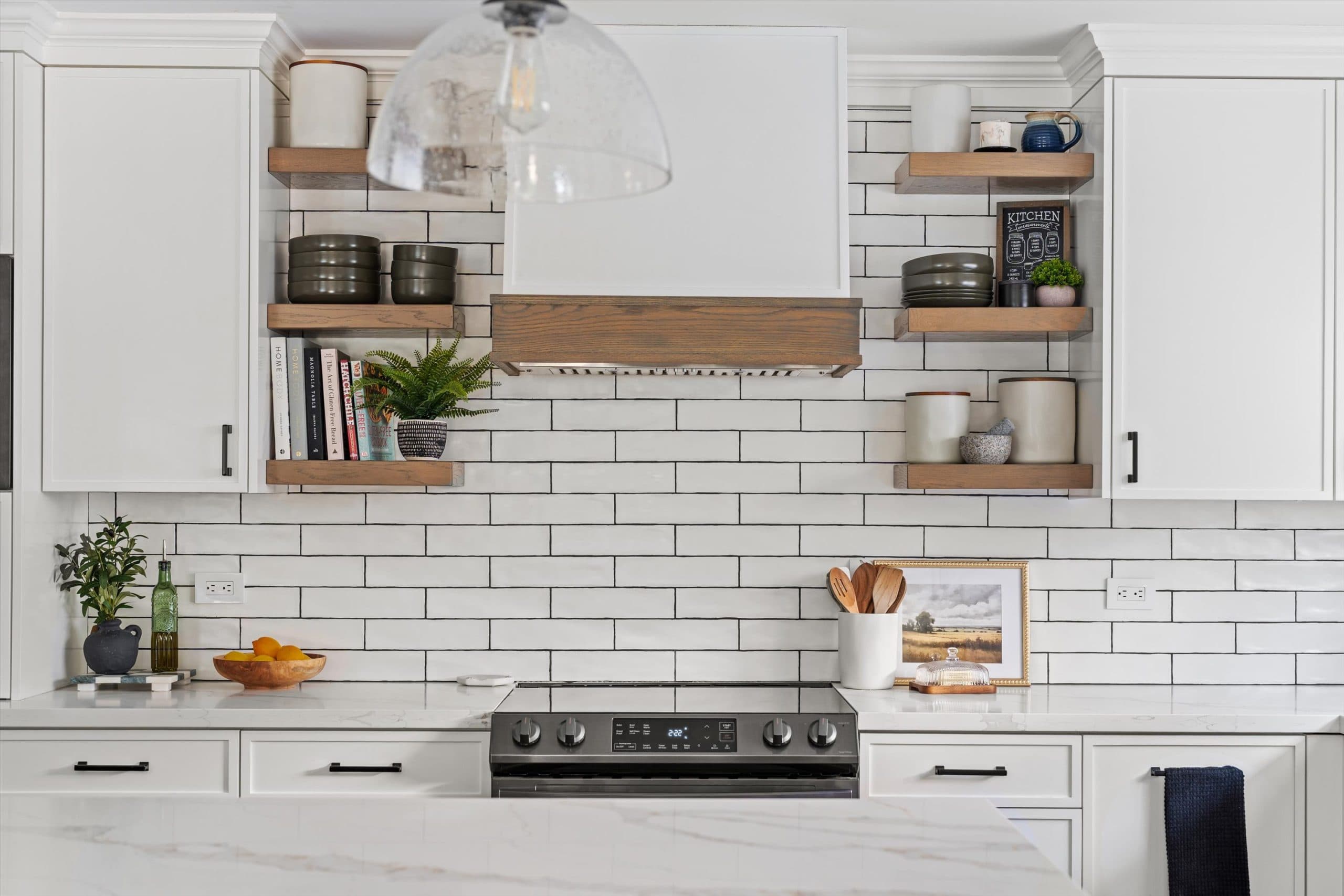 Kitchen range area with white subway tile backsplash, floating natural wood shelves displaying jars and cookbooks, stainless steel range, and white quartz countertops