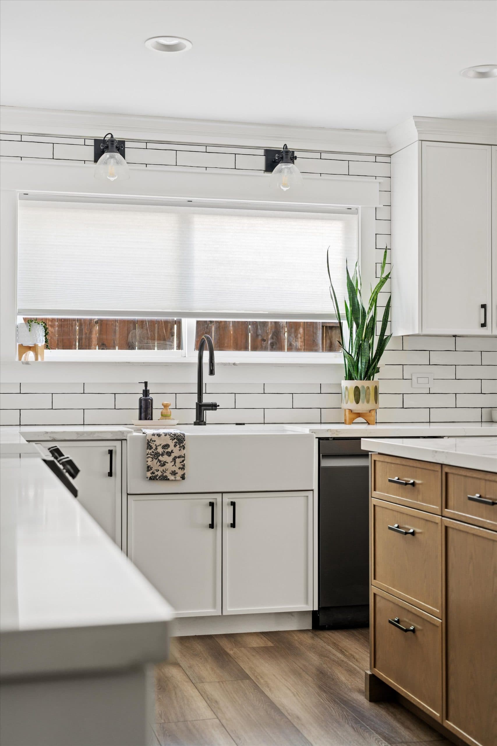 Remodeled kitchen sink area with white subway tile backsplash, black industrial sconce lighting above a large window with roller shade, white quartz countertops, and white shaker cabinetry with matte black hardware