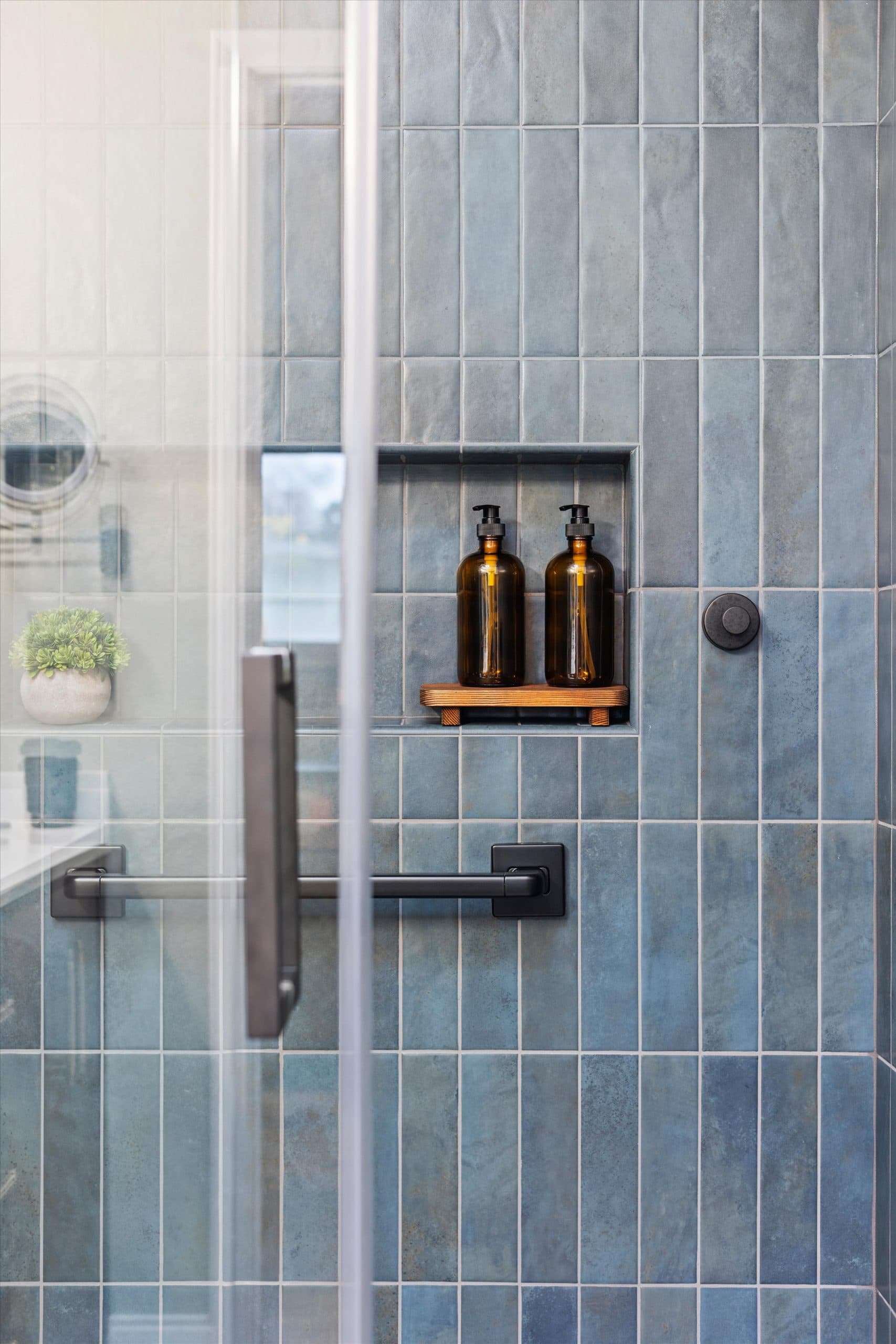 Shower niche detail showing wooden accent shelf with toiletries against blue subway tile, viewed through glass enclosure
